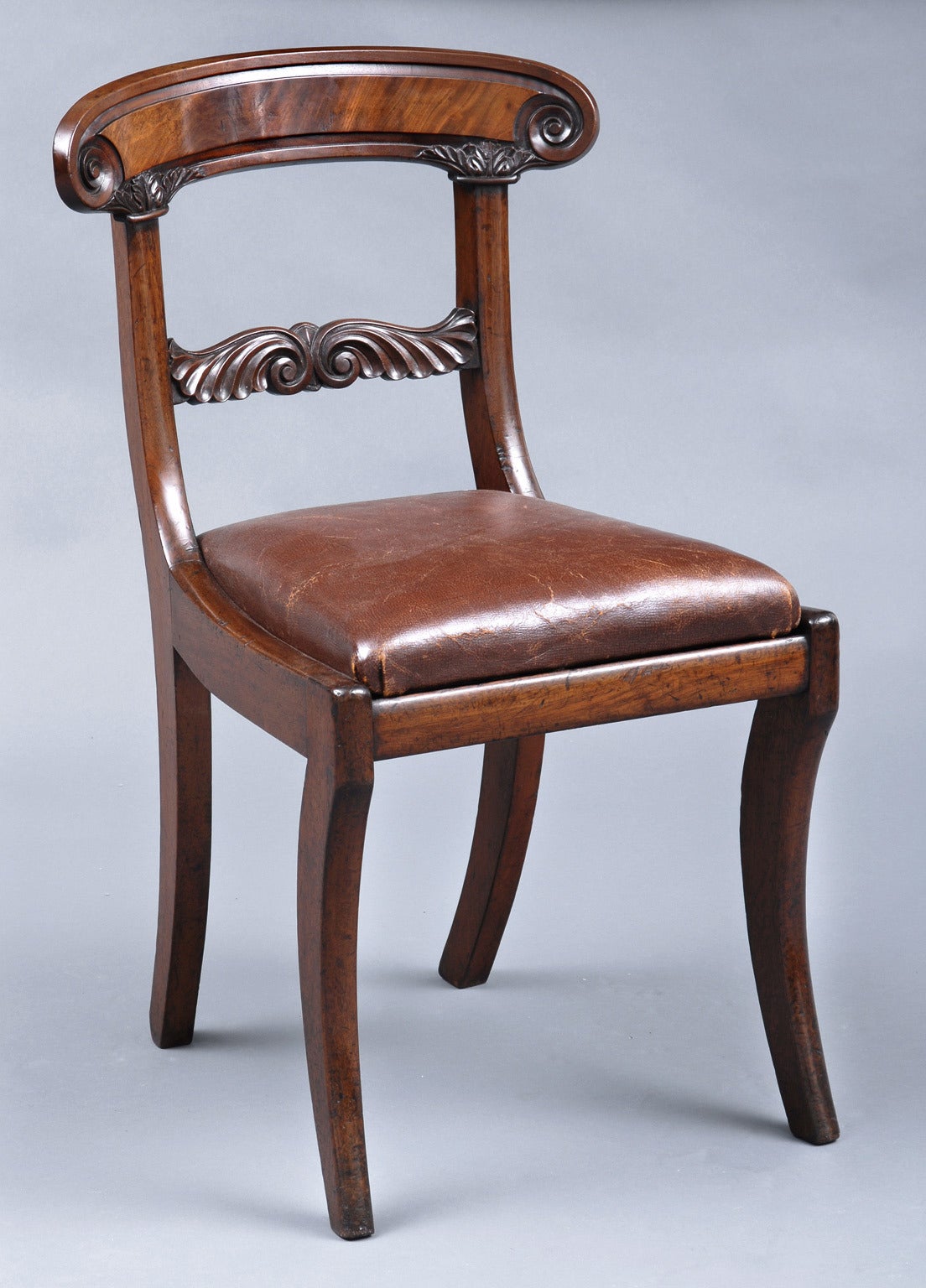 William IV mahogany side chair with curved top rail carved with scroll terminals, splat with carved leaf decoration, sabre legs, brown leather upholstered drop-in seat.

 