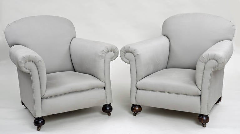 Pair of large club chairs with rounded backs, outwardly scrolled arms, tight seat, raised on beech bun feet on casters, upholstered in gray cotton fabric.