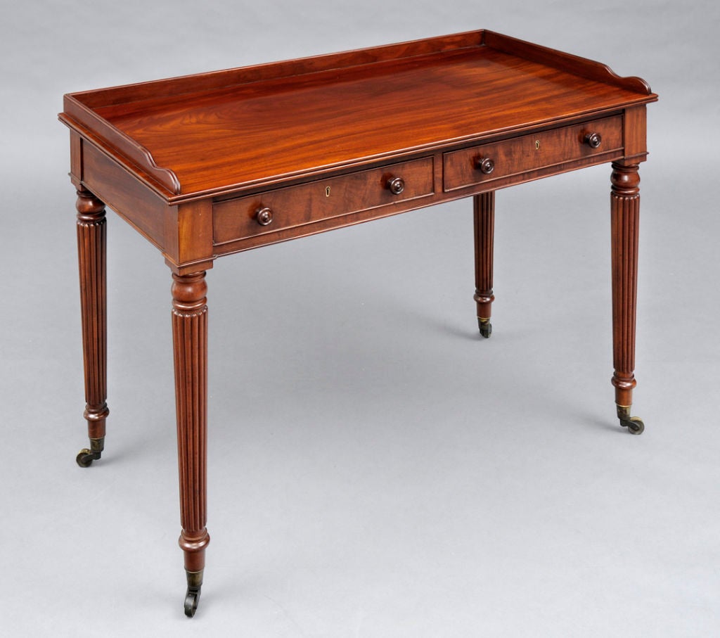 Mahogany writing table in the style of Gillows by George Oakley, London. It has two cock beaded frieze drawers with turned knobs, a shaped gallery on three sides, elegant turned and reeded legs ending in brass casters.