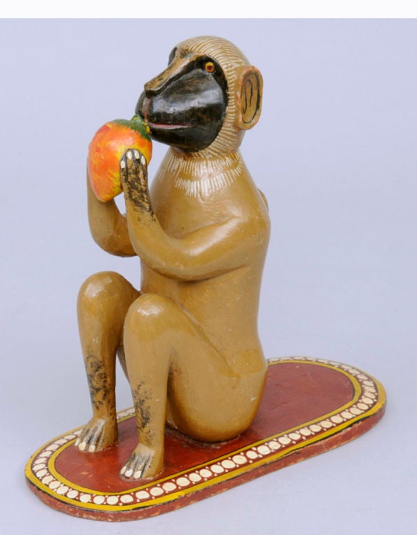 Whimsical Indian carved wood and polychromed monkey sitting on a red oval carpet with white dotted border eating a mango.