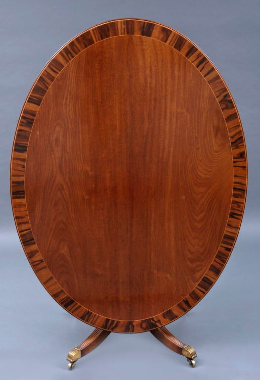 Regency oval mahogany tilt-top breakfast table with reeded top edge, top cross-banded with boldly figured coromandel wood and satinwood stringing on a turned baluster support, four out-swept reeded splay legs ending in brass terminals on casters.