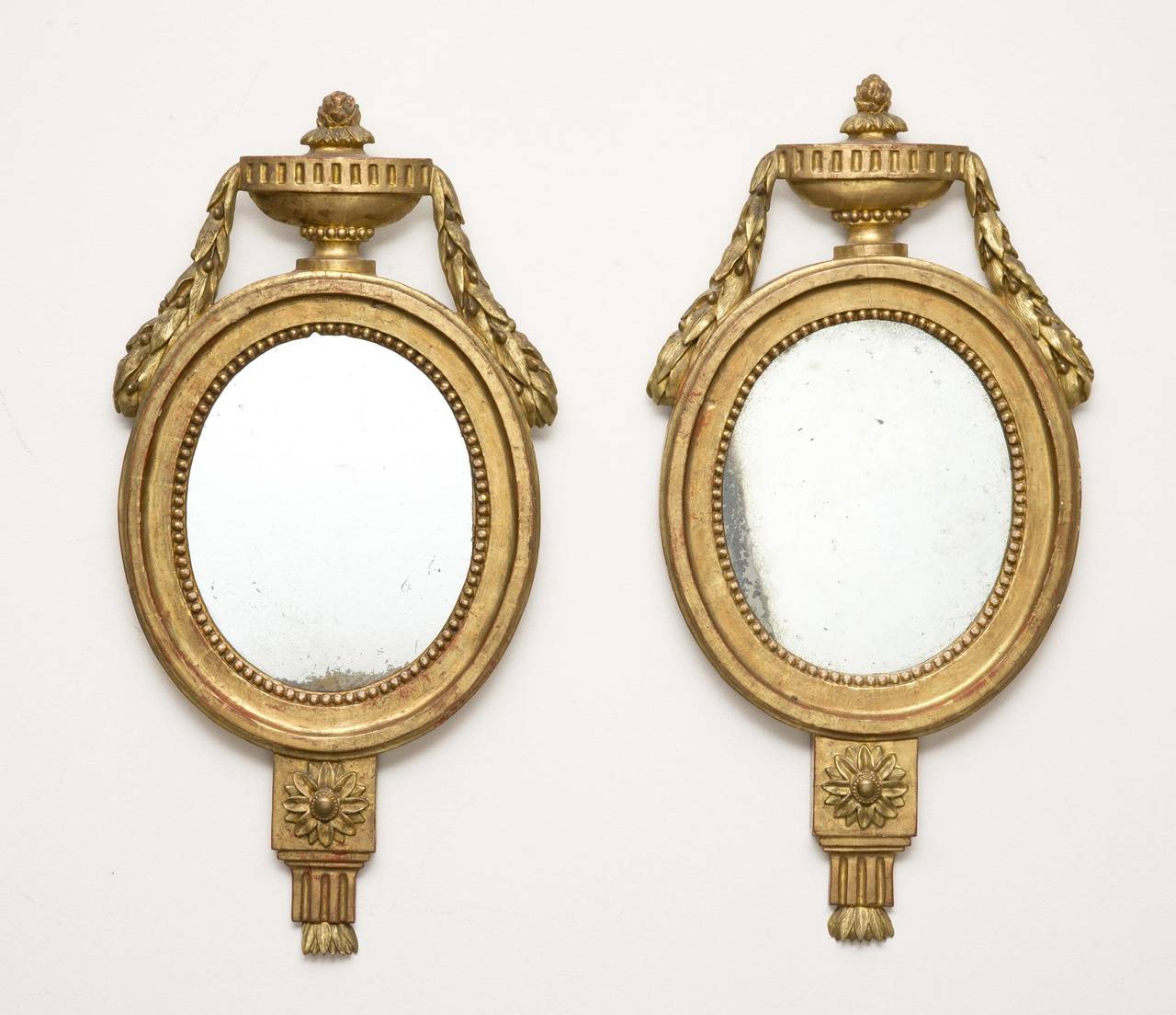 Pair of Italian giltwood oval mirrors, surmounted by a carved urn with laurel leaf swags, beaded inner molding and carved pendant below centered with a flower.

