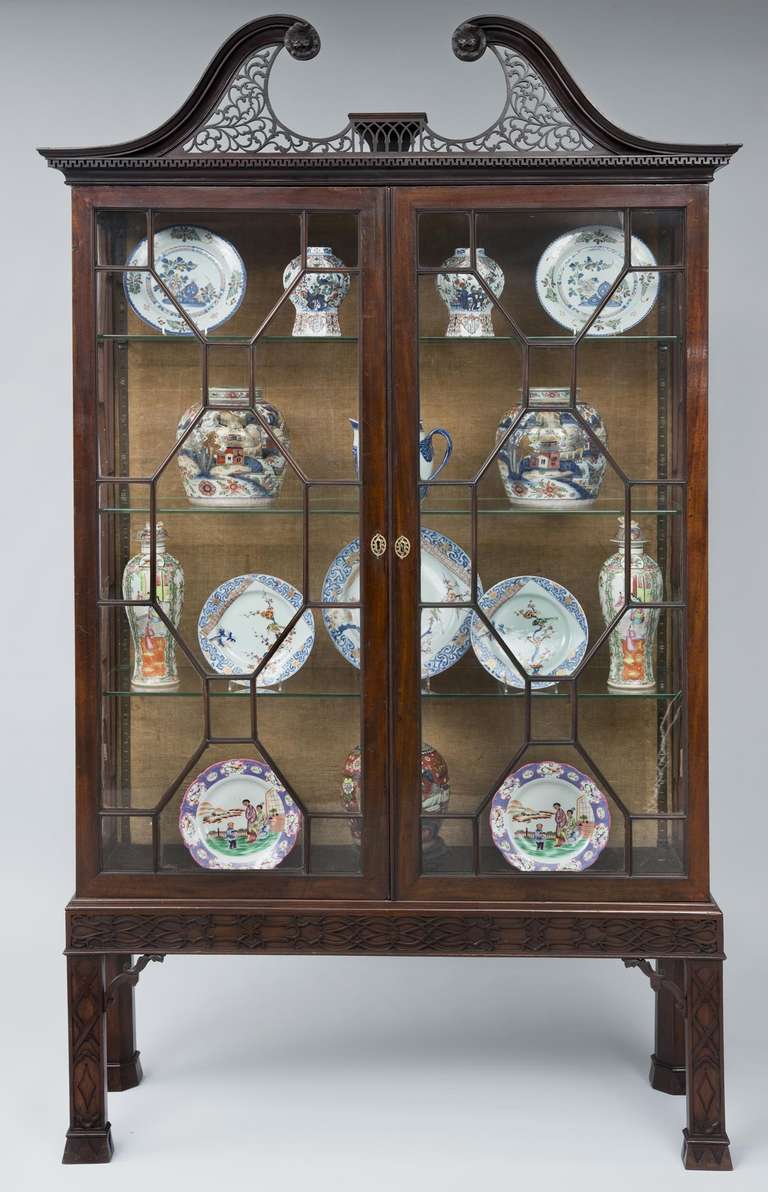 Georgian mahogany glazed display cabinet with swan neck pediment with pierced scrolling fretwork and carved roses, above Greek key molding, the doors enclosing glass shelves, raised on later Chinese Chippendale stand.  There are four glass shelves;