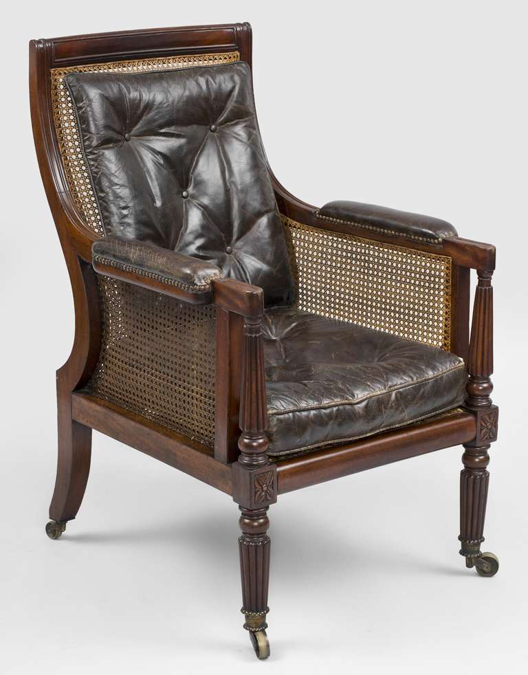 Regency mahogany library caned armchair with curved and molded back frame, padded leather arm rests with brass nailheads, reeded column arm supports above a carved four-petaled square design, the caned back and seat with loose brown buttoned leather