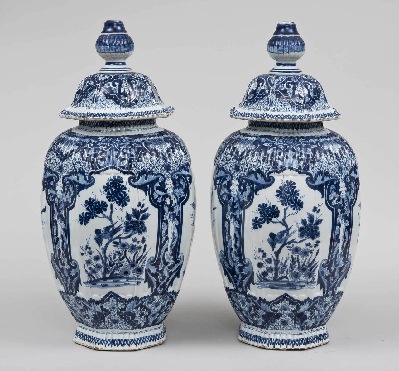 Pair of large blue and white Dutch Delft vases, the lids with bulbous shaped finials, octagonal and ribbed sides comprised of four panels decorated with flower urns, birds in trees and separated by narrow panels of putti topped by a masque. The