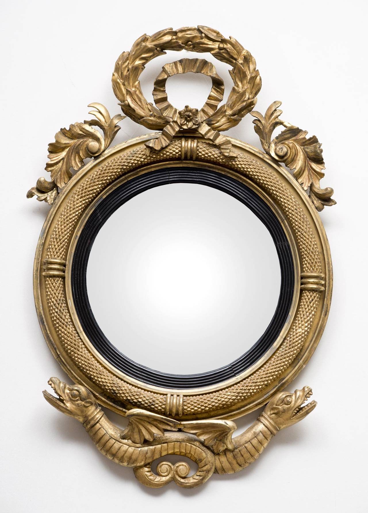Rare American Federal period giltwood convex mirror, the circular plate and ebonized reeded slip set within a boldly molded frame carved to appear like snake skin, surmounted by a carved laurel wreath and ribboned bow flanked by stylized leaves. The