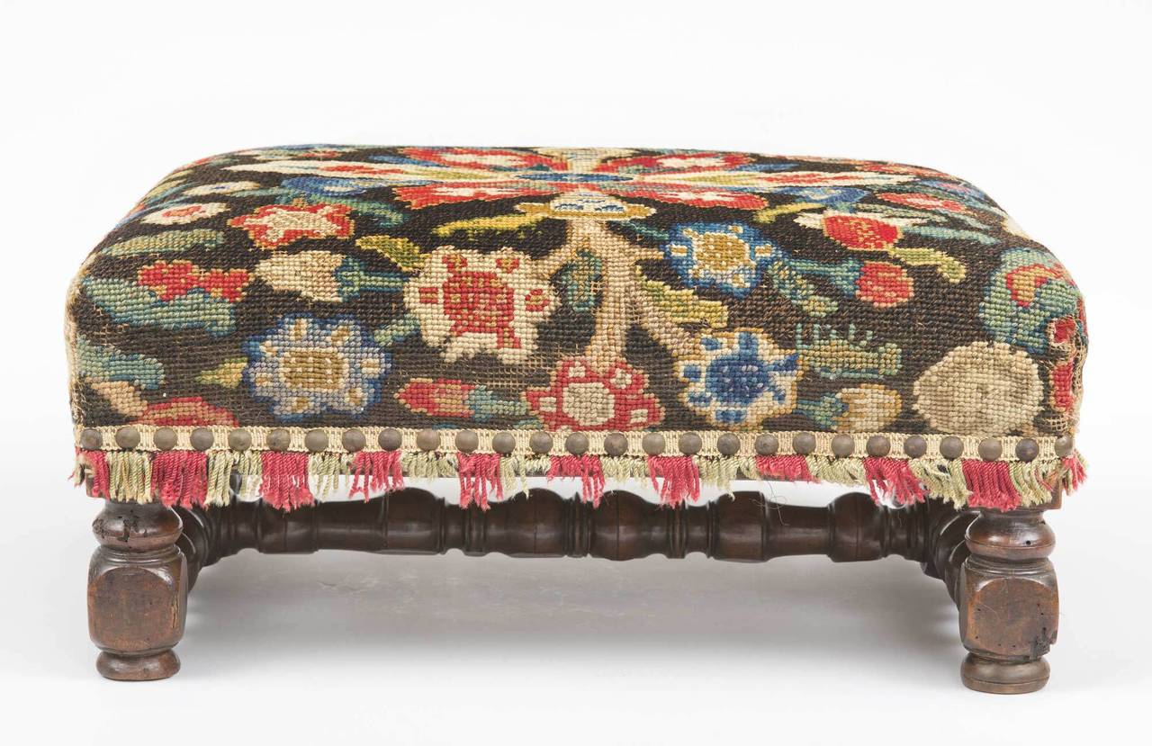 Early French walnut joined footstool with boldly turned legs and stretchers. Lovely patina. Upholstered in a colorful wool needlework in a striking floral design with fringe and brass nail heads.

          