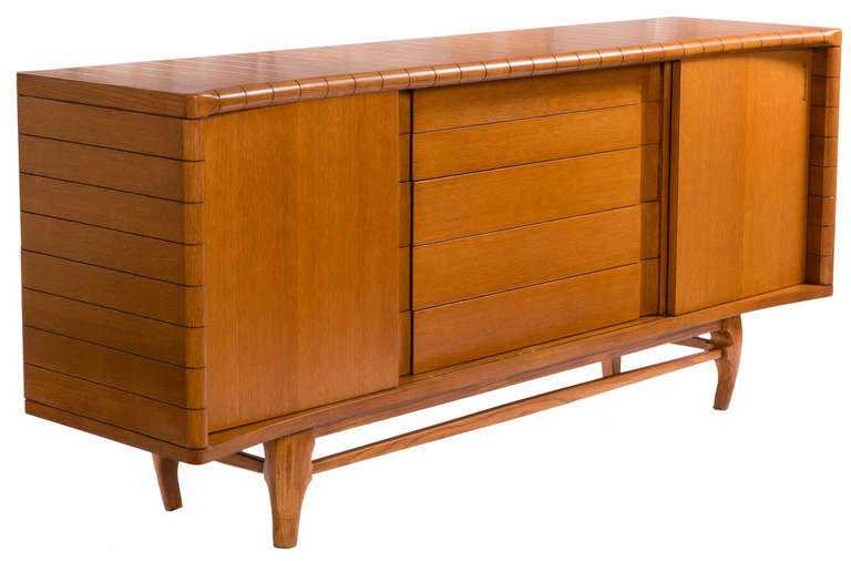 Expressive sideboard circa 1955 in solid oak with contrasting groove detail wrapping the piece, curled feet and concave front. Sliding doors reveal interior shelves while concealing the drawers, with all gaps between the moving elements aligning