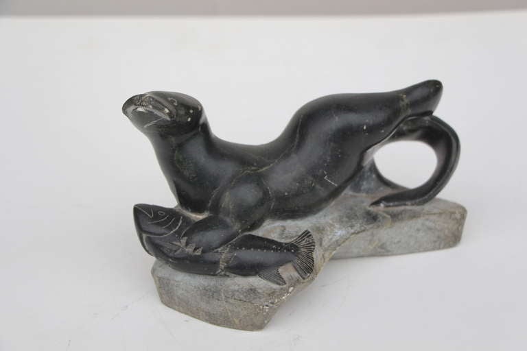 An older inuit carved soapstone whimsical sculpture of a seal or walrus catching a fish, mounted on stone. It is signed underneath. 