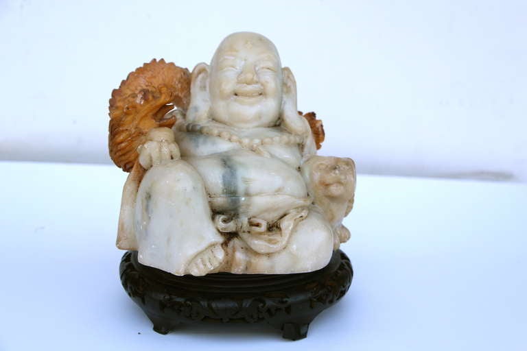 A whimsical Asian carving probably made of alabaster and partially dyed. It may also be some other softer stone. The base is not original to the sculpture, we added it. The Buddha as a dragon wrapped around his shoulders and there is cat sitting by