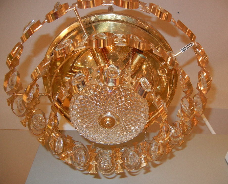 Der vergoldete Messingbaldachin trägt einen kreisförmigen Rahmen mit vertikalem Florentiner Design, der sich mit gebogenen ovalen Schleifen abwechselt, die tropfenförmige, abgeschrägte Kristallanhänger in drei absteigenden Ebenen enthalten. Die