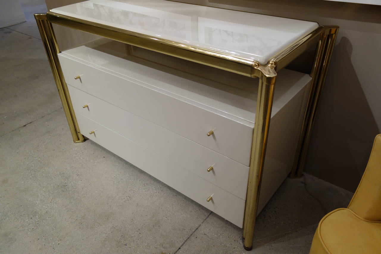 An Italian cream colored lacquered dresser within a substantial brass frame finished on all four sides featuring a floating top over a recessed space with three brass knobbed pull out drawers. A pair of companion night stands are also available.