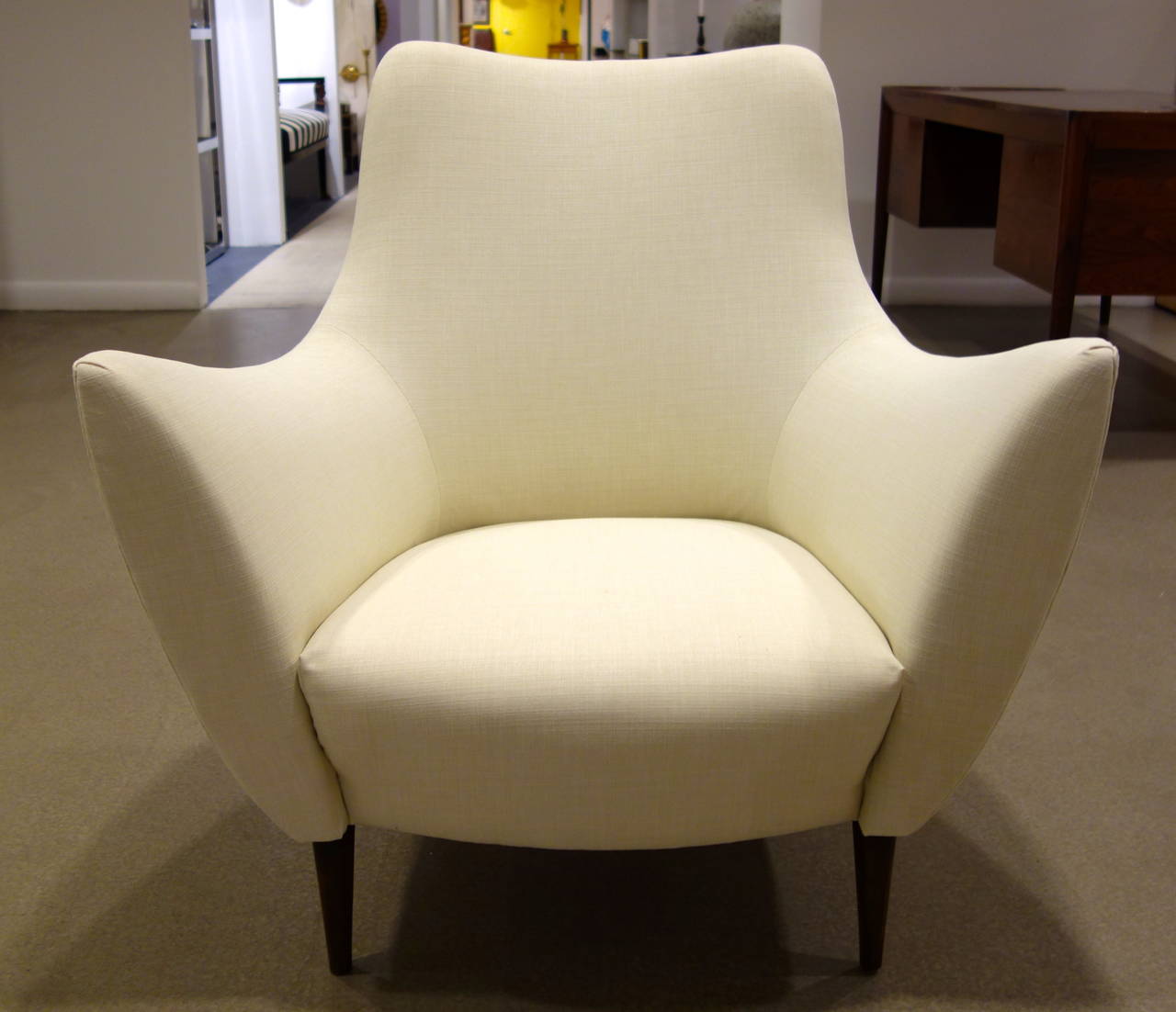 A pair of Mid-Century Style sculptural Italian lounge chairs with shaped, flared arms with tight backs and seats resting on tapered, conical wood feet newly upholstered in off-white textured fabric.  Dimensions are listed below and the arm height is