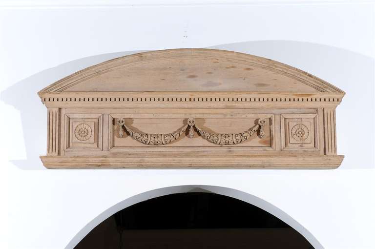 A pair of English neoclassical style carved pine overdoors with swag motifs and dentil molding from the mid 19th century. Each of this pair of English architectural elements features a curved pediment, sitting above a dentil molding. The lower