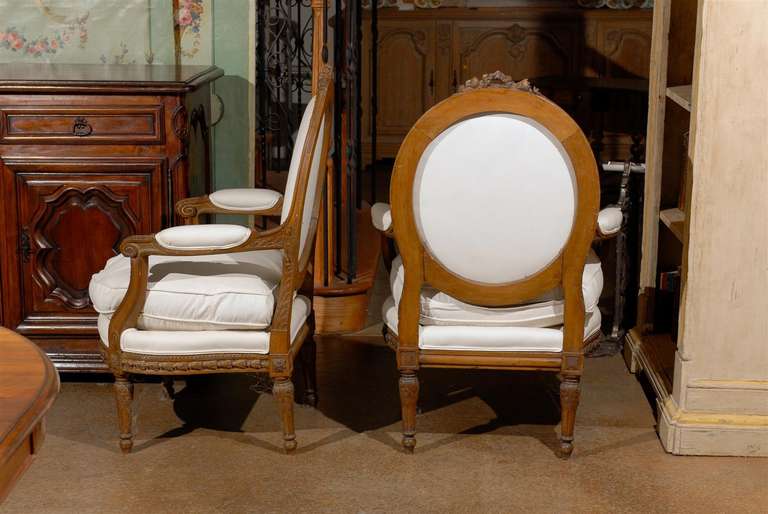 A pair of French Louis XVI style carved wooden upholstered fauteuils cabriolets with oval backs from the early 19th century. Each of this pair of French armchairs features a richly carved oval back with foliage themed crest. The back and seat are