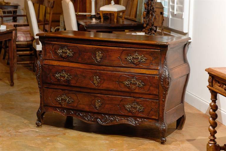 A French Louis XV period carved walnut three-drawer bombé commode with original hardware from the 1730s. This French bombé chest features a rectangular top with rounded edges over three delicately carved drawers that alternate concave and convex
