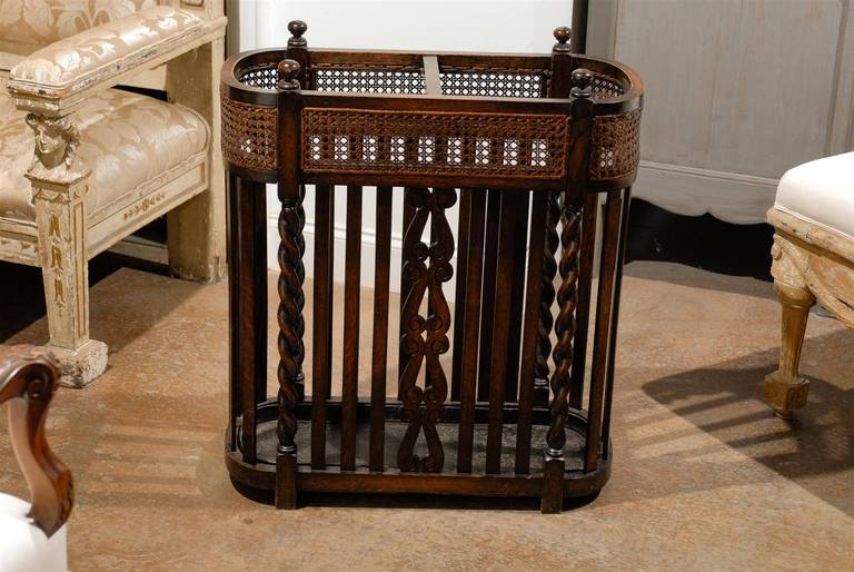 An English Victorian period wood and cane umbrella stand with metal tray inset. Born in the later years of the 19th century, this elegant English umbrella stand features an oval design, adorned with cane in its upper section. Presenting a metal tray