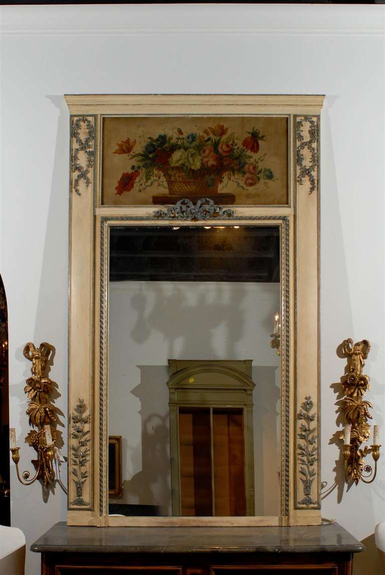 A French Louis XVI style painted and gilt trumeau mirror with floral motifs from the early 19th century. Born in the tumultuous years of the early 19th century, this French trumeau mirror features a linear cream painted frame. Two pilasters, adorned