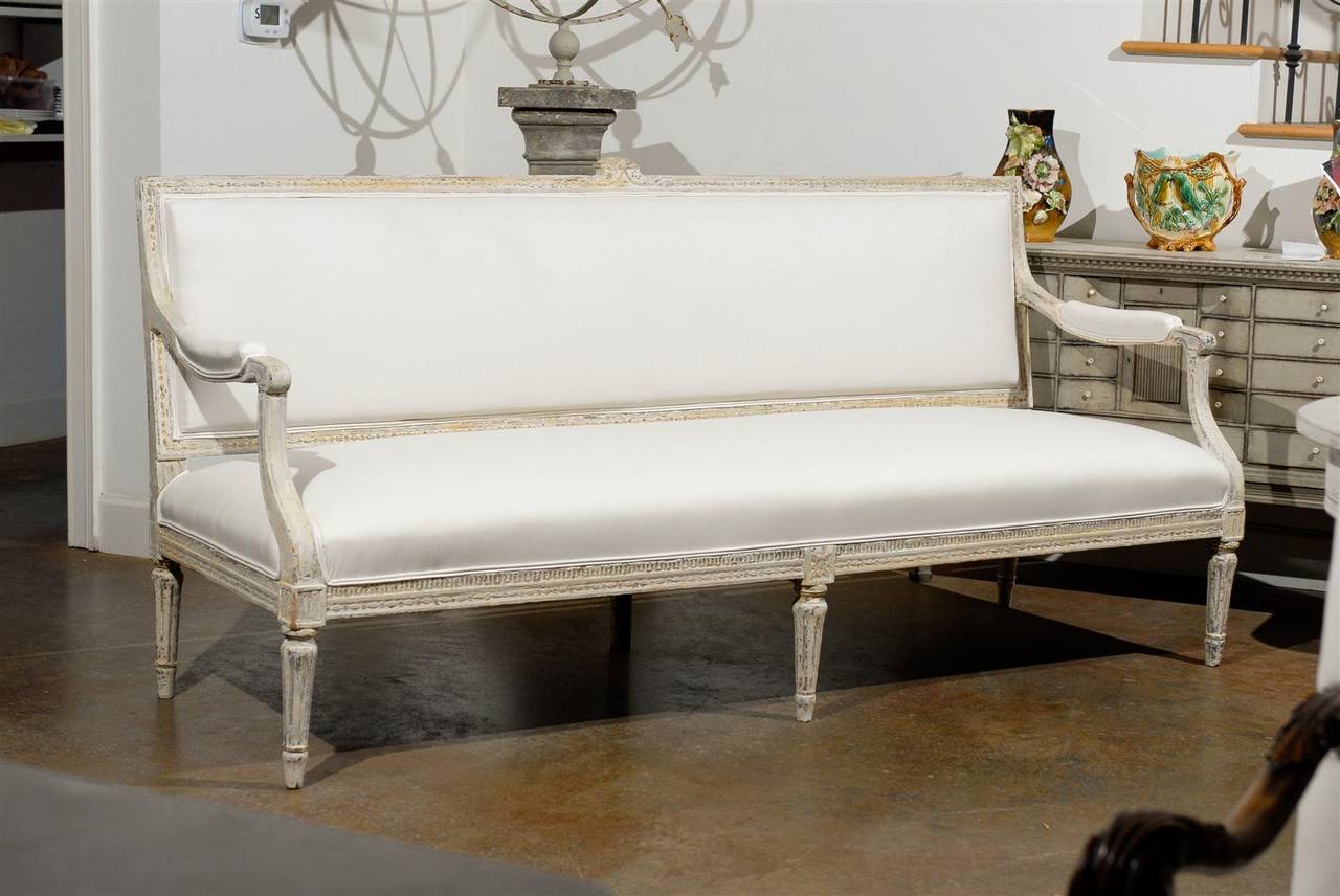 This Swedish period Gustavian painted wood sofa from the late 18th century features a slightly slanted linen upholstered back with a delicately carved crest with partially upholstered scrolled arms and deep upholstered seat. The ensemble is raised