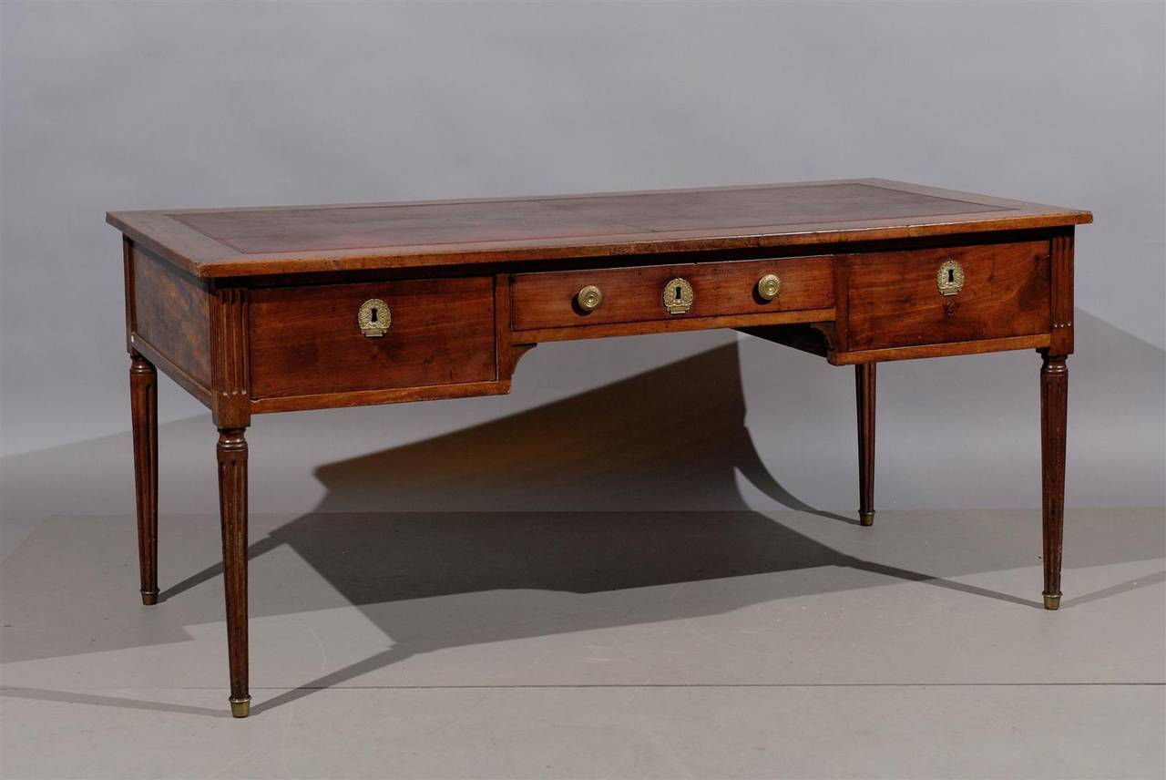 Louis XVI style walnut bureau plat with brown leather top, fluted detail on apron,  3 sliding drawers and turned fluted legs with brass caps. 

William Word Fine Antiques: Atlanta's source for antique interiors since 1956.