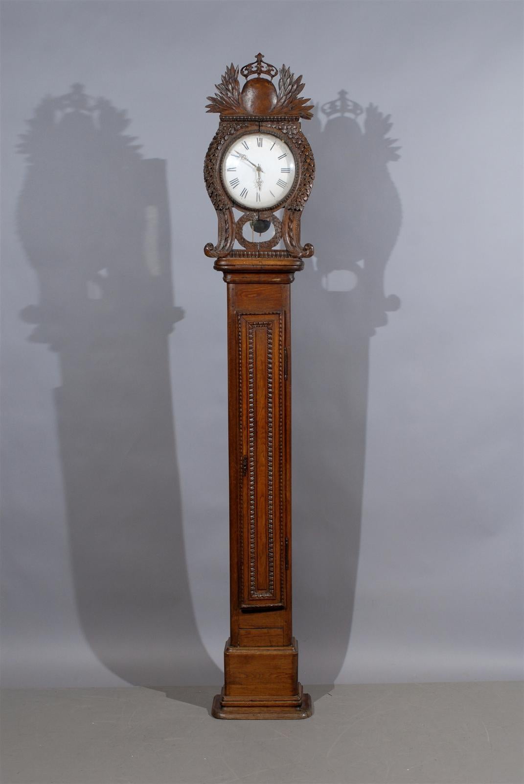 An early 19th century tall-case clock in oak with carved crest of crown and laurel leaves and round white enamel face, originating from Normandy France and dating from the turn of the 19th century. 

Clock works sold as is. Please contact us