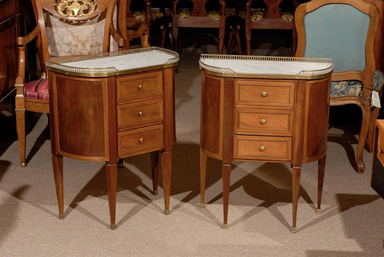 A Pair of Louis XVI Style Demilune Bedside Commodes in Walnut with White Marble Tops and Brass Galleries, dating from the 20th Century and French in Origin.

To view all of our inventory, please visit our website. williamwordantiques

William