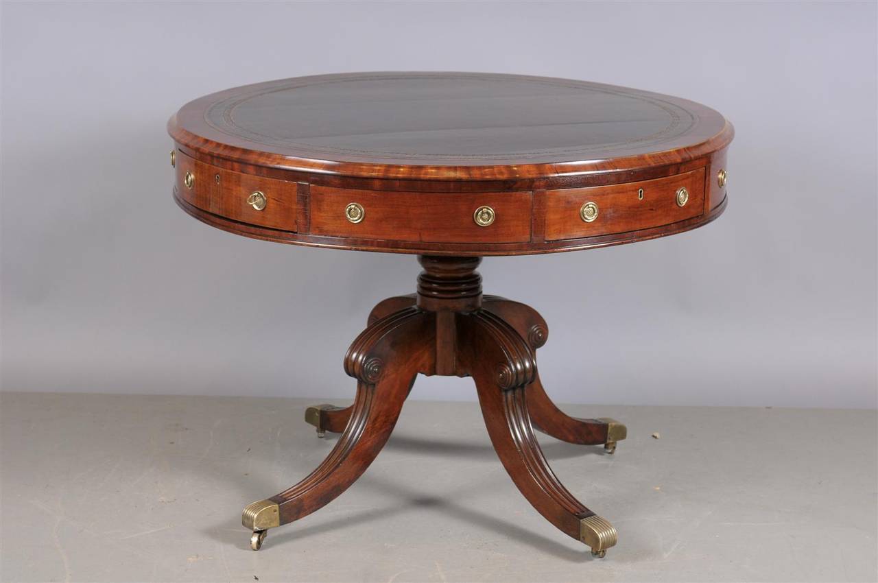 An English Mahogany drum table with brown leather top, four sliding drawers and pedestal base with brass castors. 