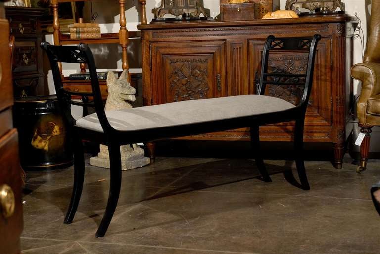 An English Empire style upholstered sleigh bench with arms from the turn of the century. This English ebonized wood sleigh bench features a rectangular seat upholstered in linen flanked by two curved fluted arms on each end with scrolling frames.