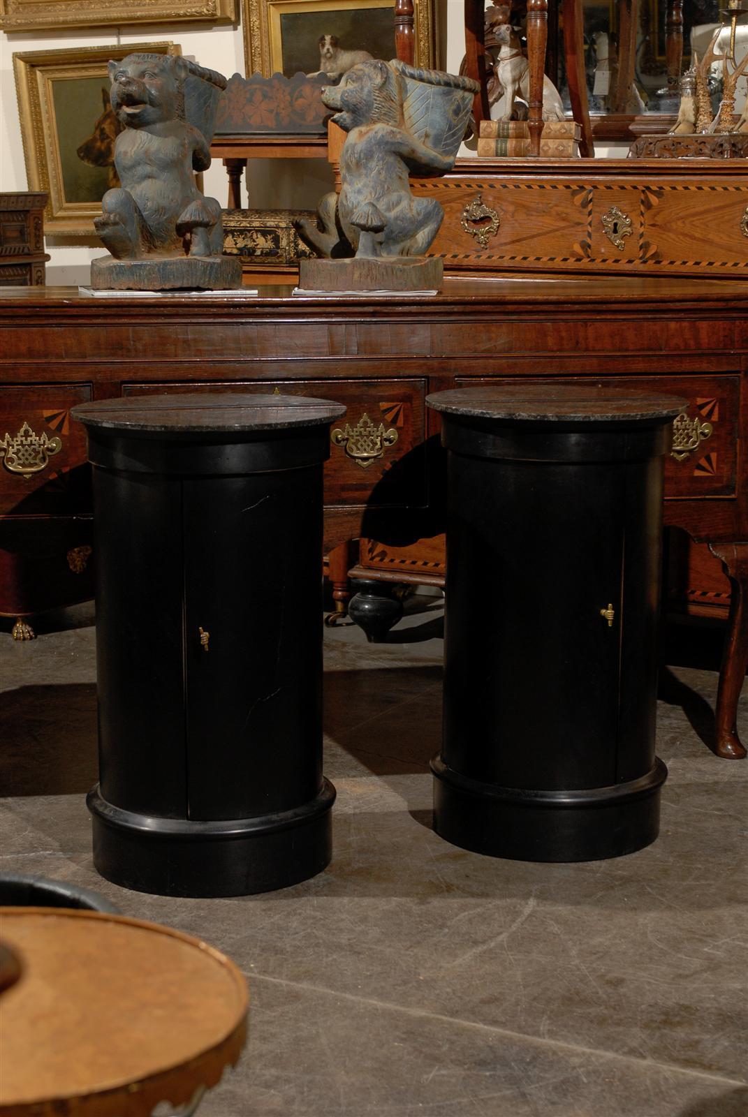 This pair of French ebonized wood column tables from the early 20th century features round dark grey or black marble tops with brown accents over circular bodies with single doors. Each door opens (in the opposite direction from one another) to