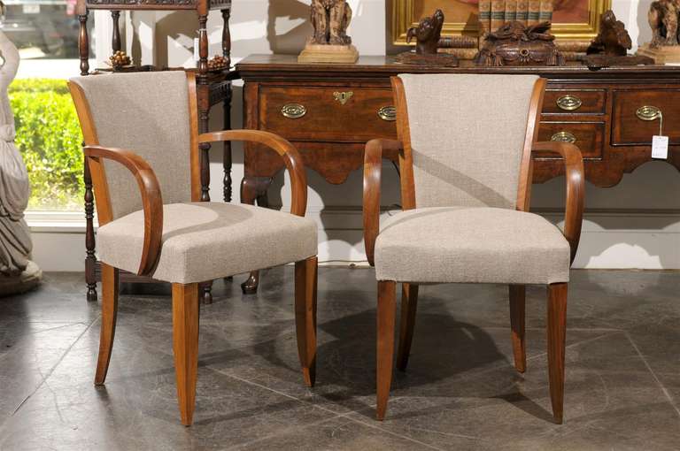 A pair of French, Art Deco walnut armchairs from the early 20th century with linen upholstered backs and seats. This pair of sculptural Art Deco armchairs, circa 1920 features a general Silhouette that favors rounded lines. The side supports on the