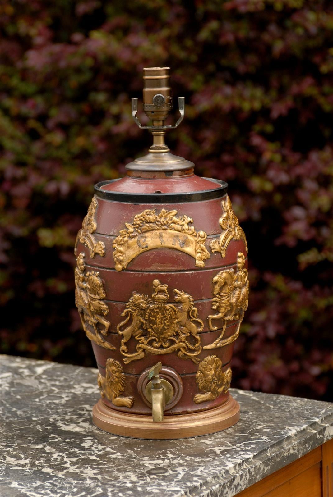An English late 19th century spirit barrel with raised gilded motifs, Gin label and brass spigot made into a table lamp. This English spirit barrel lamp features a red body, exquisitely adorned with the label Gin at the top and the United Kingdom’s