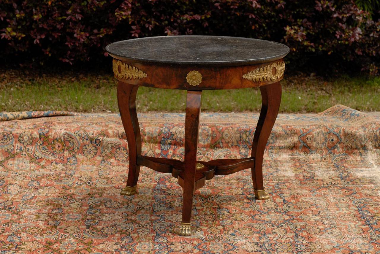 This French early 19th century Empire guéridon table features a round dark grey / black marble top over an exquisite apron, adorned with bronze floral motifs. The table is raised on three legs, showing a slight cabriole shape with gilded paw feet.