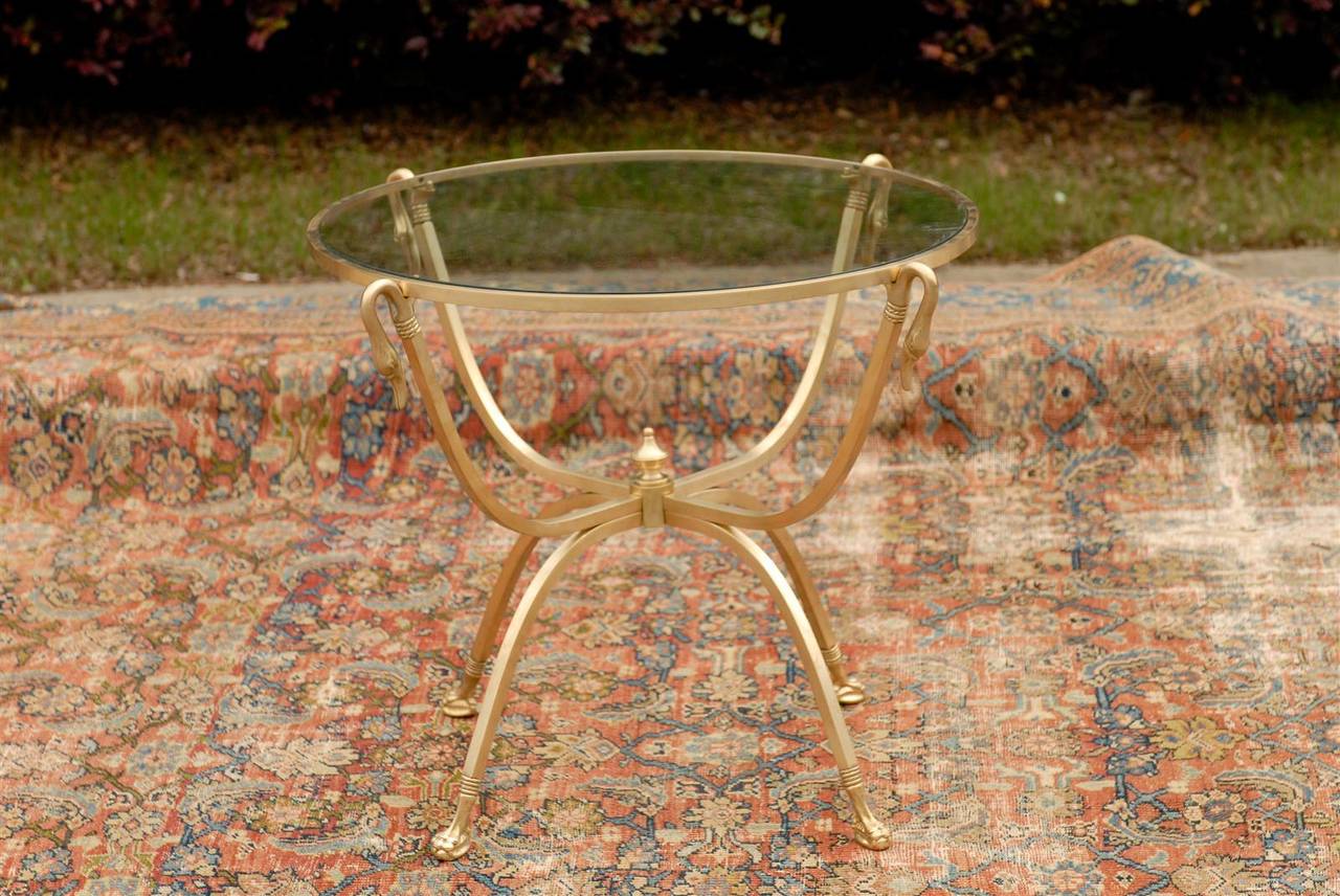 This Italian brass gueridon table from the mid-20th century features a round glass top over an exquisite gilded base. Four elegant swans support the top and gracefully converge towards the centre marked by a circular elongated finial. Curving down,
