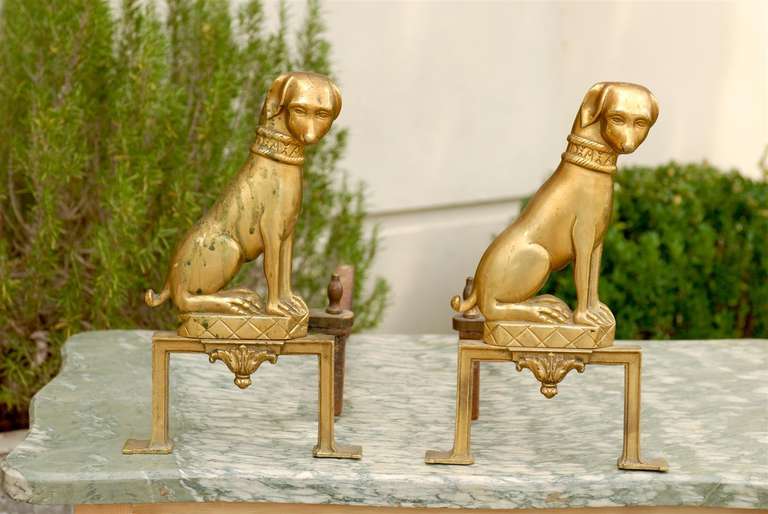 This pair of French brass andirons features two dogs quietly sitting on a rectangular base with ribbed decor and foliage motif below. These andirons are both beautiful and functional as they support the logs in a fireplace. There is a lovely