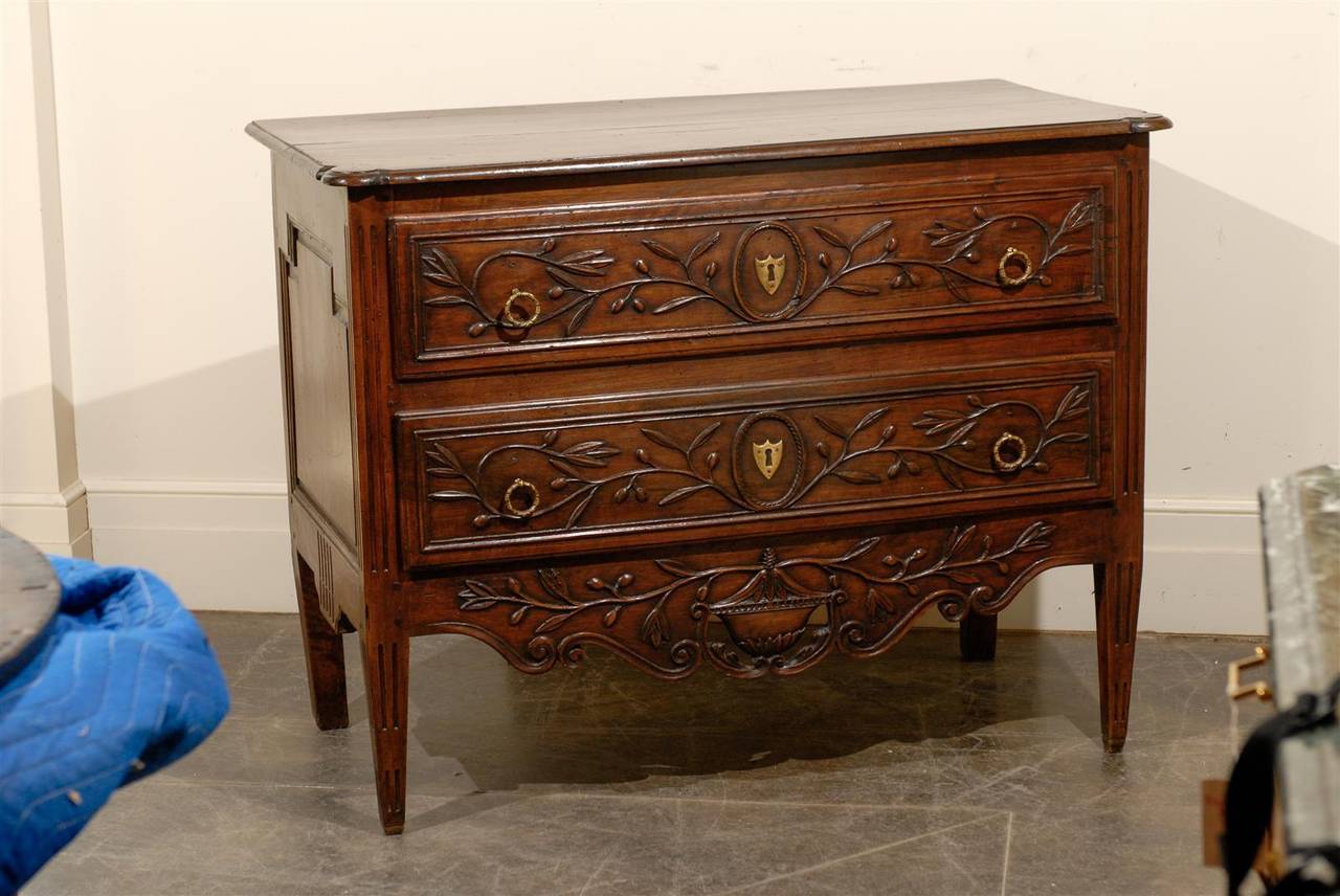 This French commode from the early 19th century features a two-plank rectangular top with carved corners over two beautifully carved and dovetailed drawers. Each drawer is adorned in its centre with a brass shield shaped escutcheon, surrounded by a