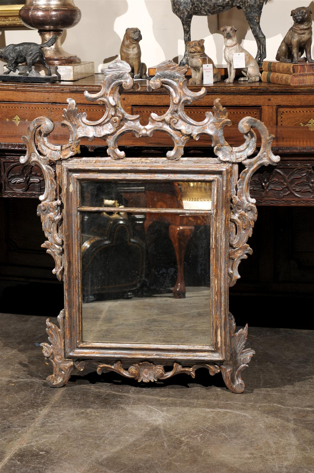 This Italian wooden mirror from the early 19th century features a Rococo style frame. The richly carved crest is made of an exquisitely refined arrangement of C-scrolls, placed symmetrically on either side of an invisible central line. The frame has
