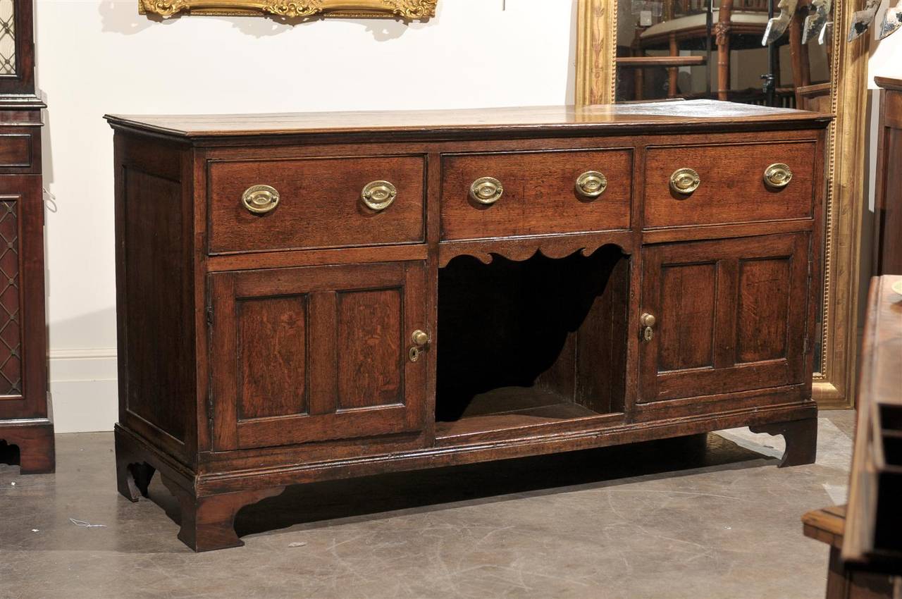 This English oak server from the early years of the 19th century features three large drawers over two doors flanking a central opening. Each drawer is adorned with two brass oval bail handles with oval backplates. The doors are decorated with