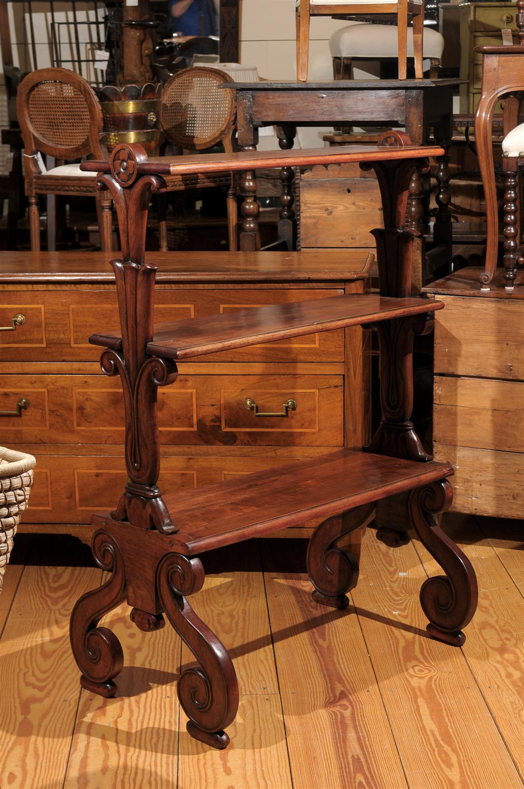 This mahogany tiered shelf from the late 19th century features three shelves over exquisite S-scroll legs. While the wooden rectangular shelves are simple in shape, the eye is immediately drawn to the lateral supports. It all starts with the volutes