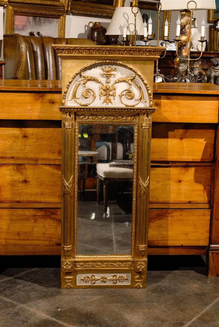 Ce miroir trumeau en bois doré français du début du XIXe siècle présente une silhouette très élancée. Le décor néoclassique de la partie supérieure est constitué de deux jolies volutes ornées chacune d'une rosace et s'enroulant autour d'une urne