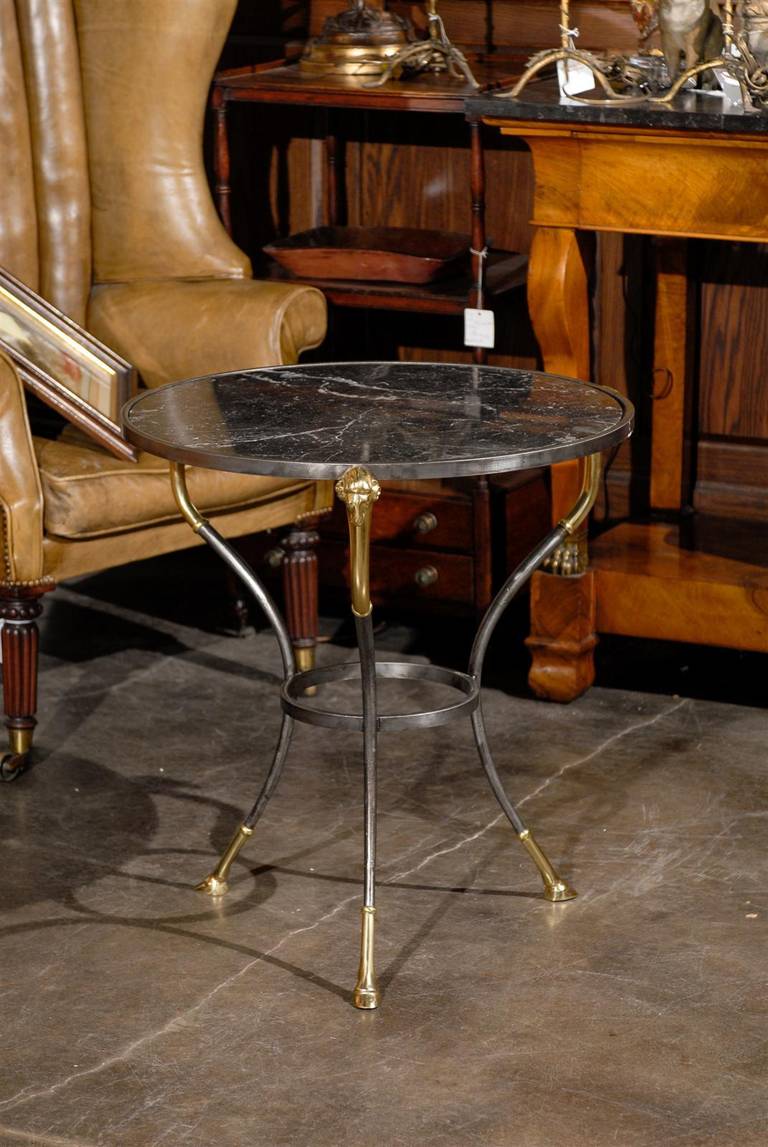 A French neoclassical style guéridon table with steel frame and marble top. This French guéridon table from the mid-20th century features a circular marble top of dark grey and white veined color, over a steel base. The tripod legs, adorned at the
