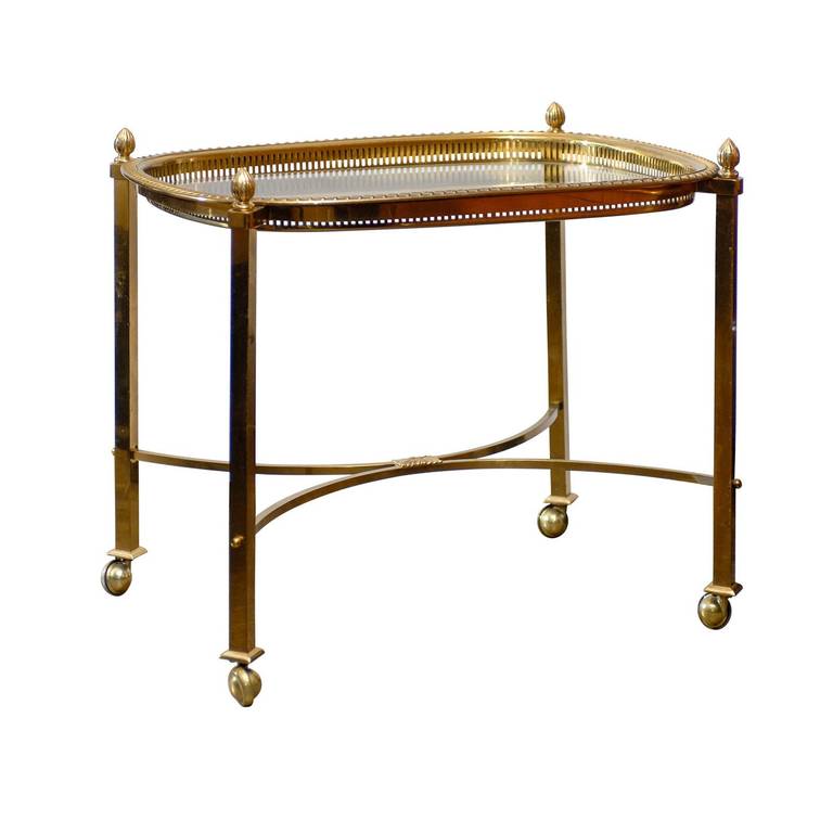A French mid 20th century brass and glass top side table. This French small size table from circa 1960 features four straight brass legs on casters each topped with a small finial, supporting a rounded rectangle tray top, made of glass. This glass
