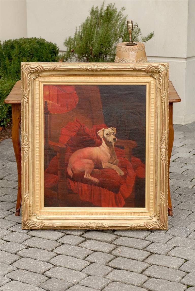 An English late 19th century oil painting of a dog sitting in an armchair with red cushion in giltwood frame. This exquisite animal painting features a short hair caramel colored dog, majestically sitting in a chair, in a manner reminiscent of