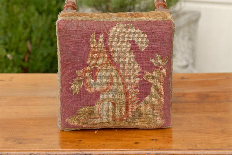 An English square shaped stool from the mid-19th century with needlepoint upholstered seat. This exquisite four legged English stool is made from yew wood, the hardest of the softwood family. The seat is covered with an adorable needlepoint squirrel