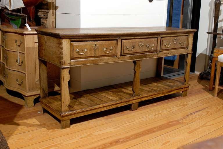 This late 18th century rustic English oak server features a rectangular molded plank top over three drawers, each with two brass swan neck handles and an open fretwork keyhole escutcheon. The server is raised on five legs, two straight in the back