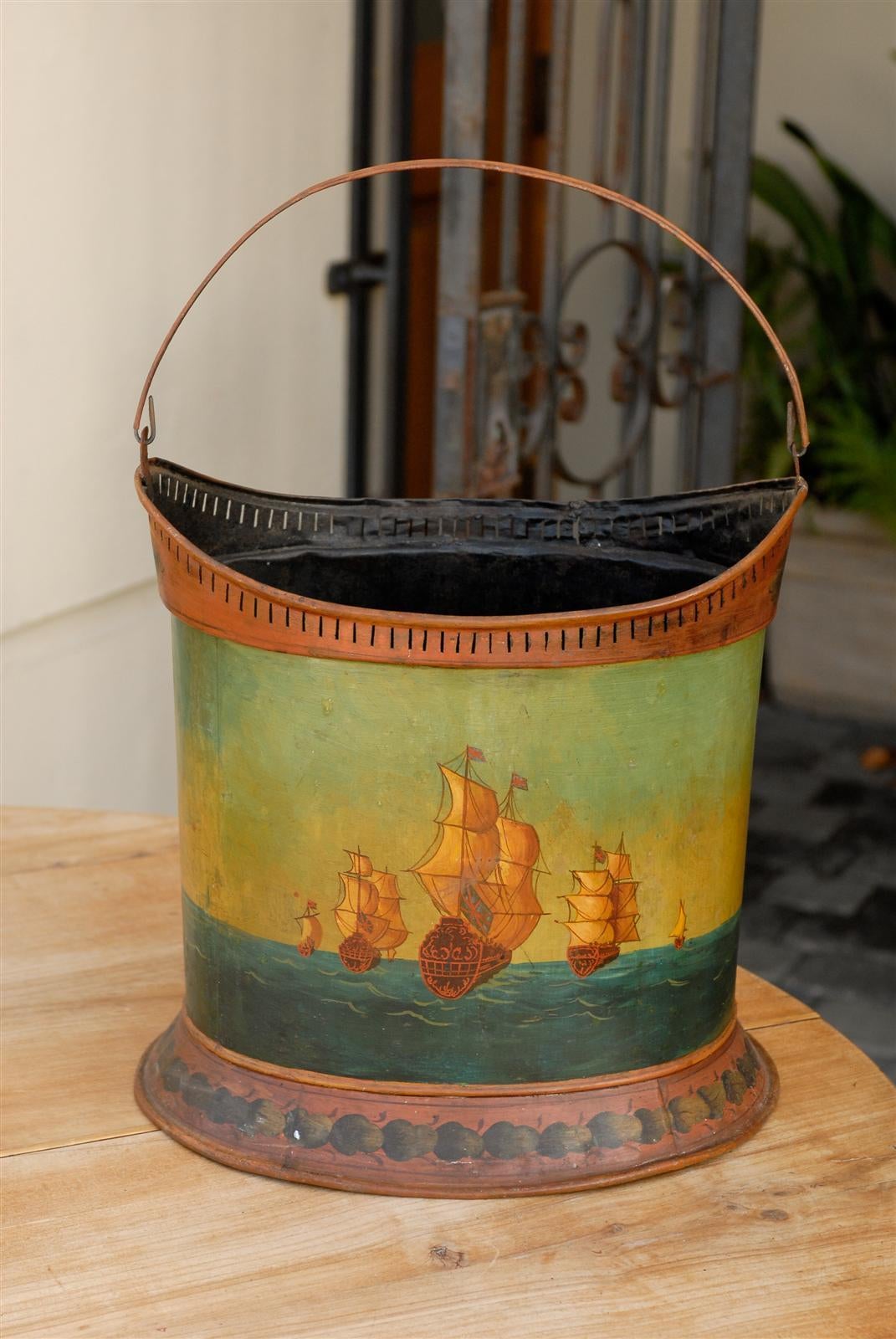 An English painted tole bucket with seascape scene and thin handle from the late 19th century. This English tole bucket features a painted scene on its body, depicting a few two-masted ships flying the Union Jack. The charming Naïve style fits the