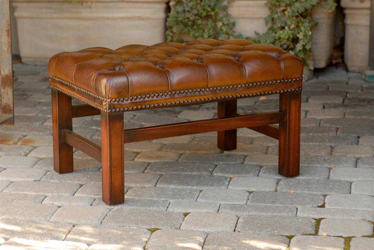 This English wooden bench from the mid-20th century features a rectangular brown leather tufted seat with double nailhead trim on the surround. The bench is raised on four simple straight legs with side stretchers placed at varying heights. This
