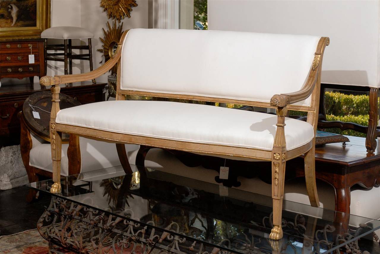 This Italian settee from the turn of the century, features an exquisite scrolled, upholstered back, connected to two arms, and adorned with stylized foliage motifs. The smooth muslin upholstered seat rests upon two saber back legs and two tapered