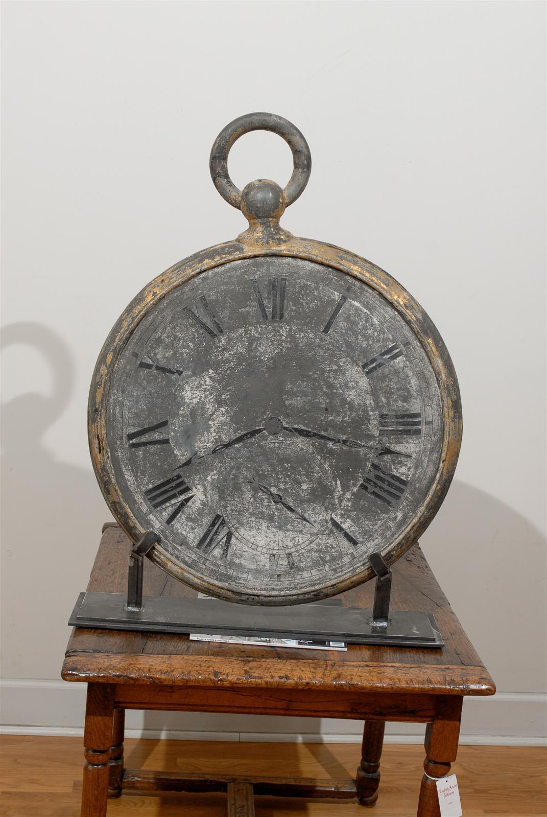 Dieses französische Großuhrenschild aus der Mitte des 19. Jahrhunderts zeigt eine überdimensionale Taschenuhr mit einer runden Schleife an der Spitze. Das graue Zifferblatt aus Zink ist mit römischen Ziffern verziert, wobei die Zeiger 8:17 anzeigen,