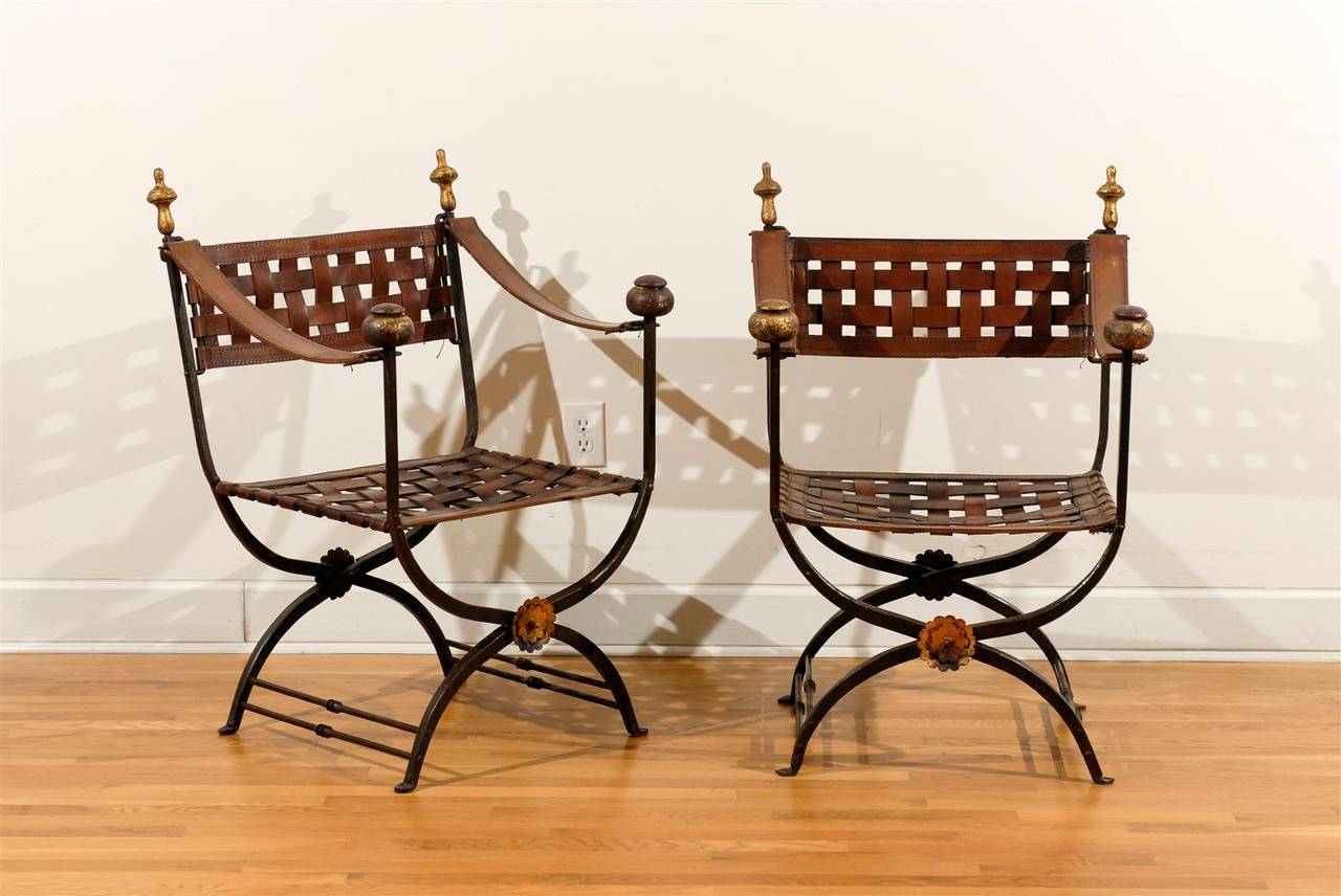 This pair of Italian Campaign Savonarola chairs from the mid-20th century features iron and brass frames with woven brown leather seats and backs. The X-shaped legs are adorned at their junction with a gilded medallion, while the upper section is