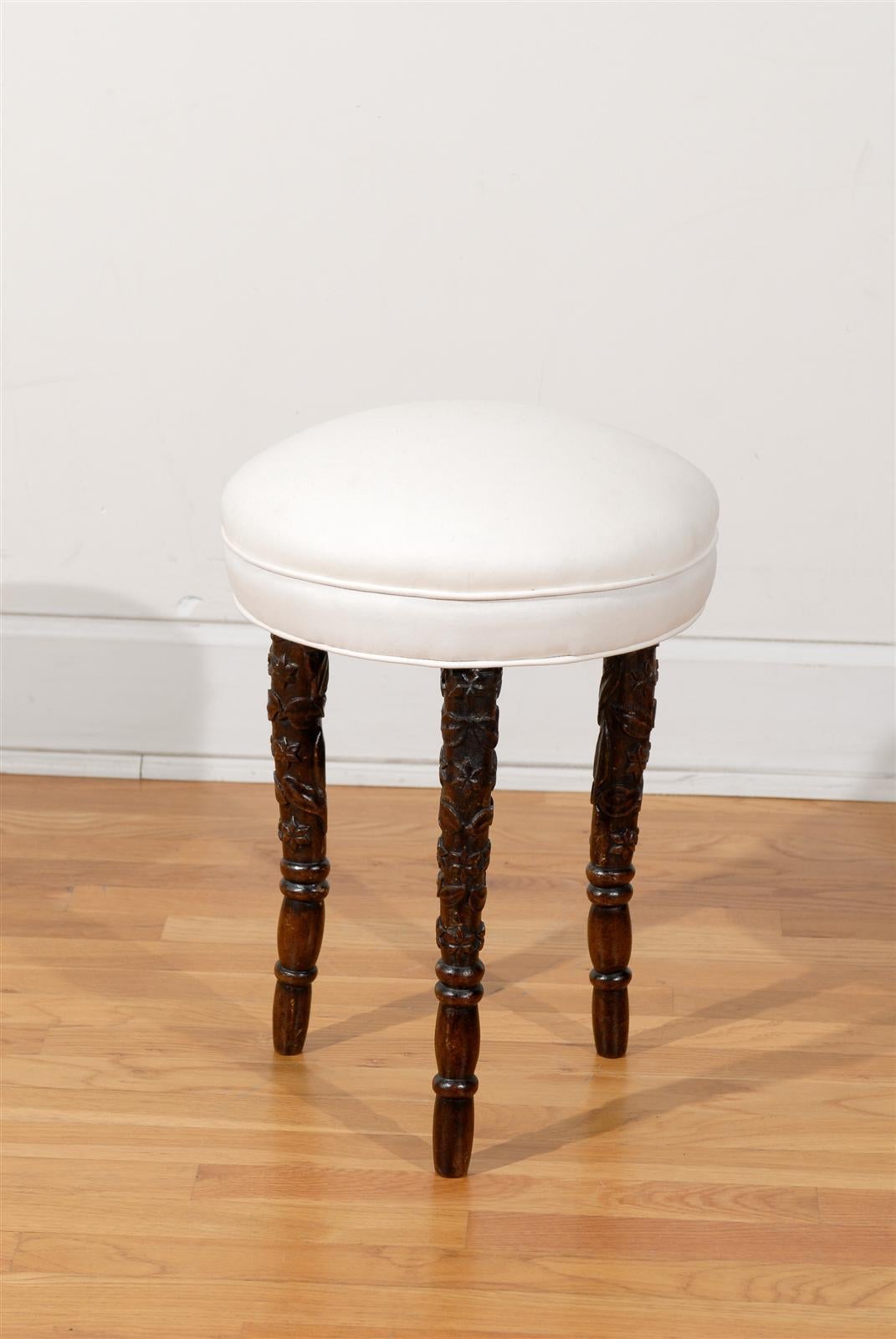 A German Black Forest carved wooden stool from the late 19th century with newly upholstered seat. This Black Forest stool features a simple single welt muslin covered round seat raised on three beautifully carved dark wooden legs. Each leg is